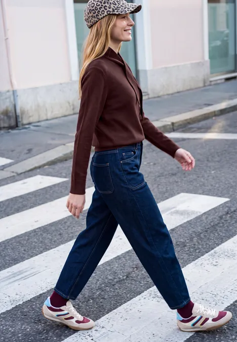 Vrouw met pet, rood jasje, blauwe jeans en sneakers steekt een zebrapad over.