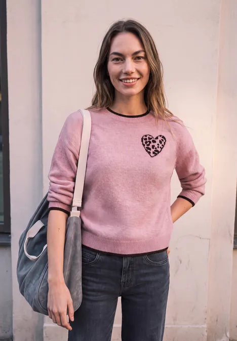 Eine lächelnde Frau in rosa Sweatshirt mit Herzprint trägt eine Tasche und lehnt an einer Hauswand.