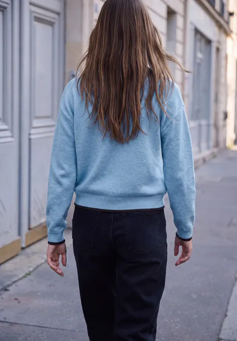 Rückenansicht einer Person mit langem Haar, hellblauem Pullover und dunkler Jeans auf einem Gehweg vor Häusern.