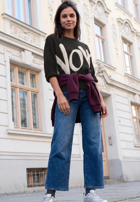 Pullover mit großem Wording Artwork Black Eine junge Frau in Jeans und Pullover mit Aufdruck 'NOW' steht lässig vor einem Gebäude.