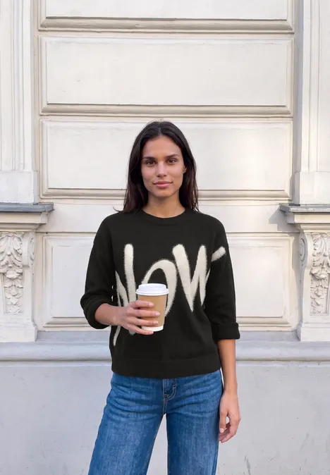 Pullover mit großem Wording Artwork Black Frau mit LOVE-Pullover hält einen Kaffee vor einer hellen Hausfassade.
