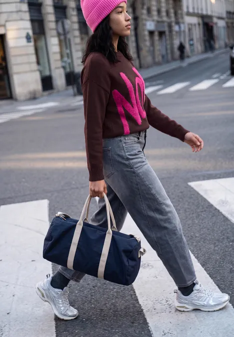 Frau mit rosa Mütze und Pullover überquert Straße, trägt blaue Reisetasche und Sneakers.