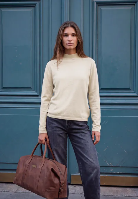 Frau steht vor einer blauen Tür, trägt cremefarbenen Pullover, dunkle Jeans und hält eine braune Reisetasche.
