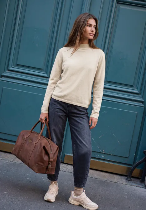 Frau in beigem Pullover, dunklen Hosen und Sneakers mit brauner Reisetasche vor blauem Tor.