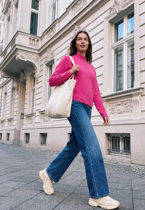 Eine Frau in pinkem Pullover und Jeans spaziert mit einer Stofftasche an einer Stadthausfassade vorbei.