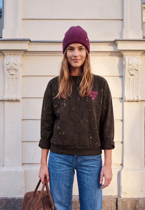Sweat-shirt ample à imprimé serpent charred brown Junge Frau mit lila Mütze, schwarzem Pullover und Jeans steht vor hellem Gebäude und hält eine Tasche.