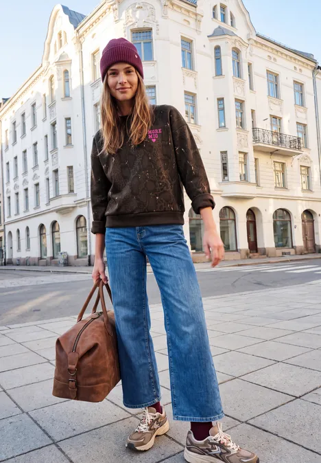 Sweat-shirt ample à imprimé serpent charred brown Junge Frau mit lila Mütze, weiter Jeans und Metallic-Sneakern posiert vor weißem Altbau, hält Reisetasche.