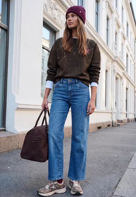 Sweat-shirt ample à imprimé serpent charred brown Eine junge Frau mit Mütze, braunem Pullover, Jeans und Tasche steht auf einem Bürgersteig vor Häuserfassaden.