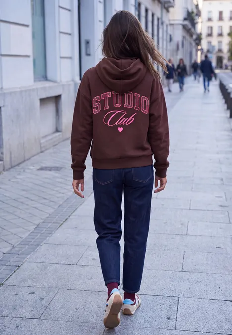 Eine Person in braunem 'STUDIO Club'-Hoodie und Jeans läuft auf einer Fußgängerzone, unscharfe Menschen im Hintergrund.