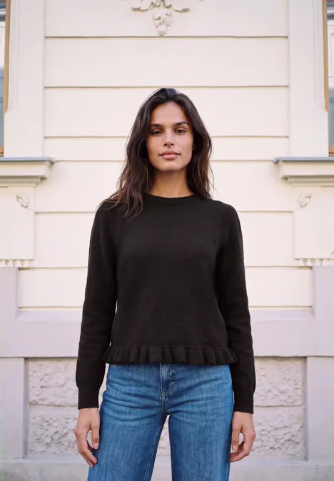 Pullover mit Rundhals und Rüschen am Saum charred brown Junge Frau vor heller Hausfassade, trägt schwarzen Pullover und blaue Jeans, schaut in die Kamera.