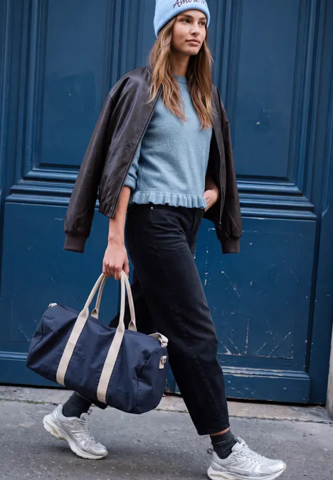 Pullover mit Rundhals und Rüschen am Saum loom blue mel. Frau mit Mütze, Pullover und Lederjacke über den Schultern, trägt Reisetasche und geht vor blauem Tor.