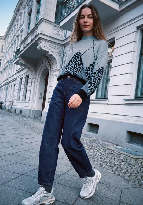 Junge Frau in weitem Cardigan, Jeanshose und Turnschuhen spaziert vor klassischer Häuserfassade.