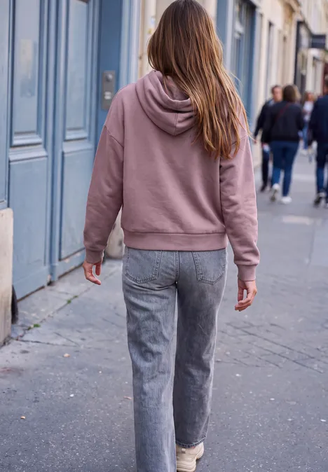 Sweat à capuche ample hazy rose Eine Person in rosa Kapuzenpullover und grauen Hosen geht auf einem Bürgersteig in der Stadt.
