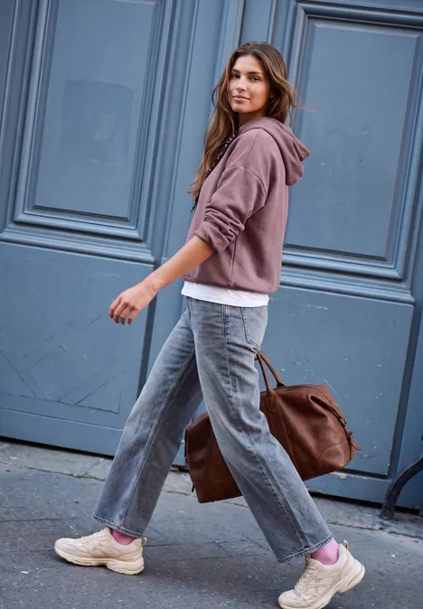 Sweat à capuche ample hazy rose Eine junge Frau in Hoodie und Jeans trägt eine große Tasche und geht an einer blauen Tür vorbei.