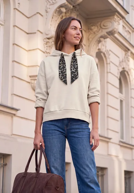 Junge Frau in Hoodie und Jeans trägt braune Ledertasche vor klassischer Hausfassade.