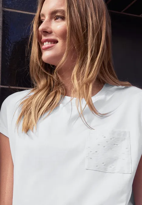 Frau trägt weißes T-Shirt mit Brusttasche vor dunklem Hintergrund.