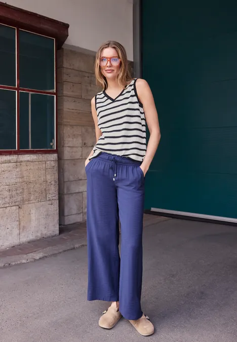 Eine Frau in gestreiftem Top und blauen, weiten Hosen steht lässig mit Händen in den Taschen vor einer Wand.