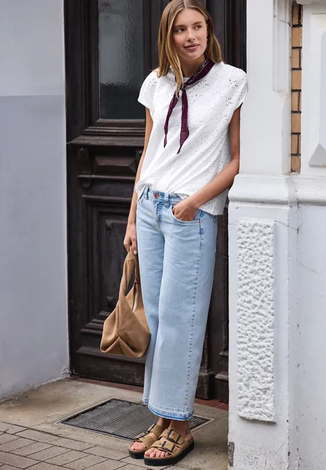 Persoon staat bij een gebouwingang, draagt een witte blouse, lichtblauwe spijkerbroek, sjaal, sandalen en bruine tas.