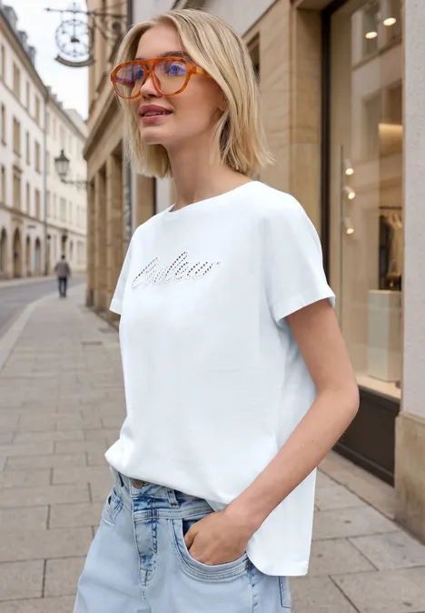 Eine Frau in weißem T-Shirt und Jeans steht mit der Hand in der Hosentasche auf einer Stadtstraße.