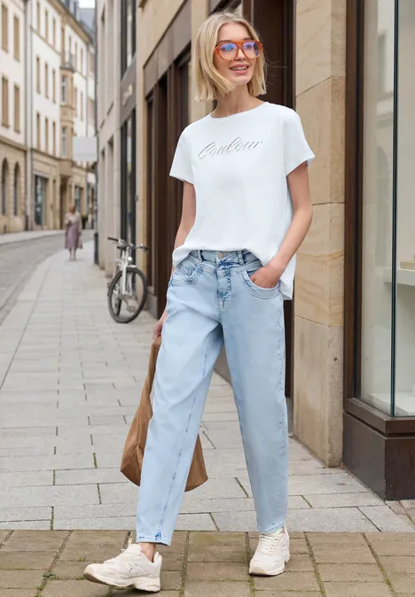 Junge Frau in weißem T-Shirt und hellblauen Jeans lehnt lässig an einer Hauswand in einer Altstadtgasse.