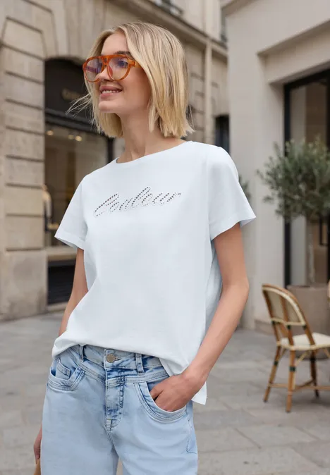 Eine Frau steht entspannt in der Stadt, trägt Jeans und ein weißes T-Shirt mit dem Aufdruck 'Couture'.