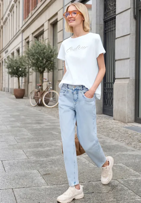 Junge Frau in Jeans, weißem T-Shirt und Sneakers geht entspannt über eine Stadtstraße.