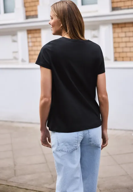 Een vrouw van achteren in een zwart T-shirt en lichtblauwe spijkerbroek staat op de stoep voor een huis.