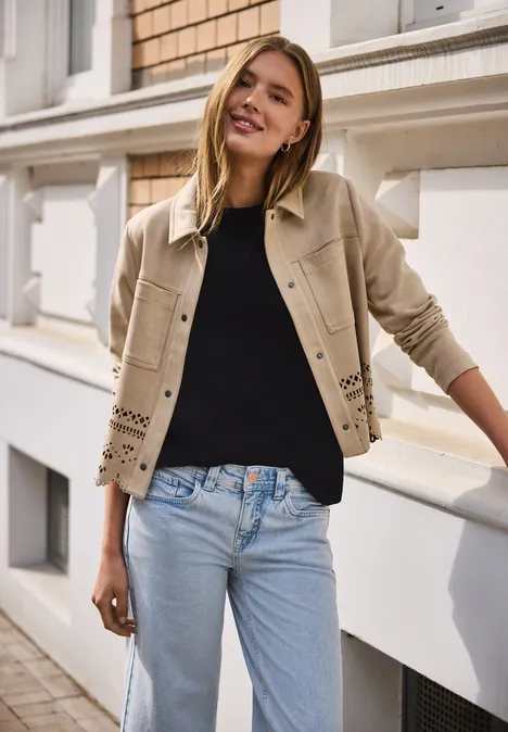 Vrouw leunt tegen gevel, draagt beige jas, zwarte trui en lichtblauwe jeans