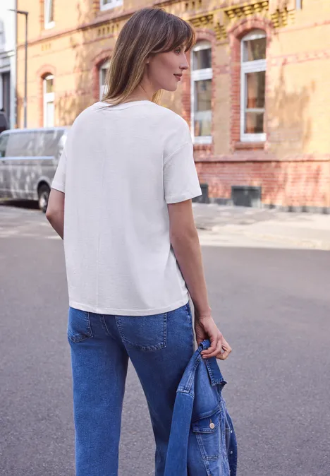 Shirt met ronde hals en tekstopdruk White Een vrouw in jeans en wit T-shirt staat met een spijkerjasje in de hand op straat voor bakstenen gebouwen.