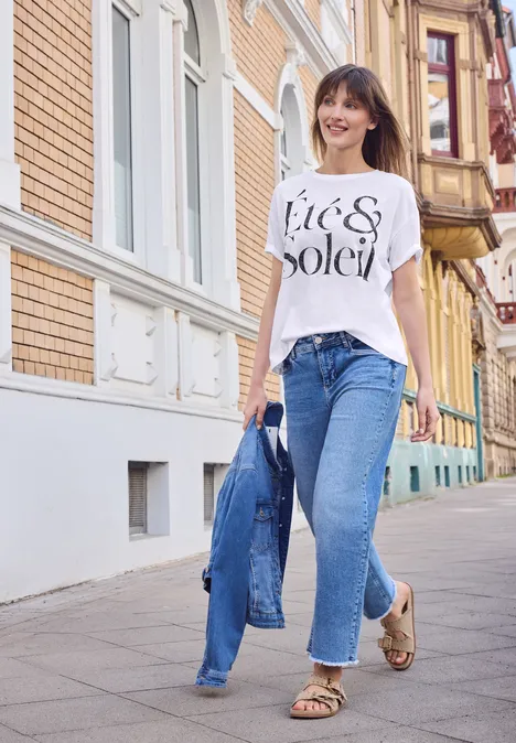 Shirt met ronde hals en tekstopdruk White Vrouw in spijkerbroek, t-shirt en sandalen loopt met spijkerjasje in de hand over een zonnige straat.