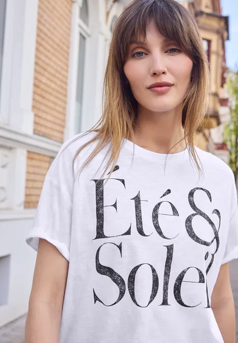 Shirt met ronde hals en tekstopdruk White Jonge vrouw draagt een wit T-shirt met zwarte tekst 'Été & Soleil' voor een licht bakstenen gebouw.
