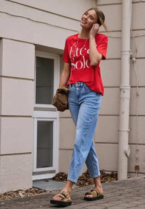 Frau in rotem Shirt und Jeans, trägt Sandalen und hält einen Hut vor Hausfassade