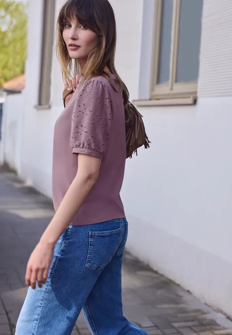 Frau in rosa Pullover mit Spitzenärmeln und blauer Jeans geht draußen an Hausfassade vorbei.