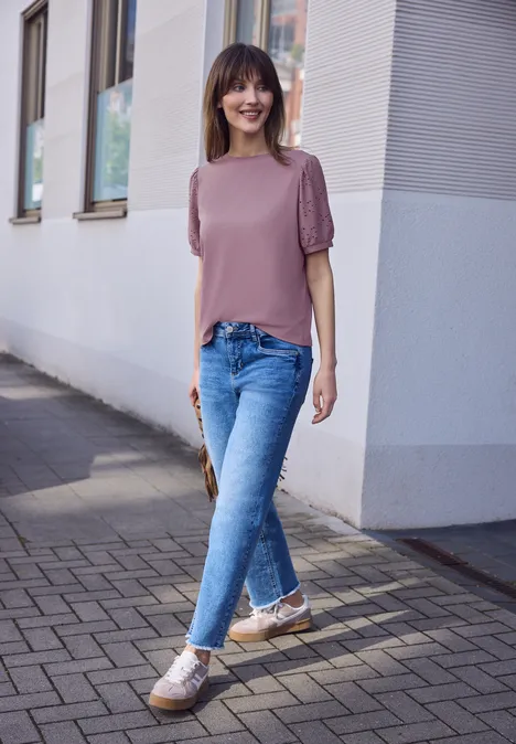 Eine junge Frau in rosa Oberteil mit Puffärmeln, blauen Jeans und Sneakern geht auf einem Gehweg an Häusern vorbei.