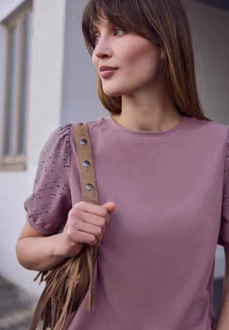 Junge Frau steht vor Gebäude, trägt hellviolettes T-Shirt mit Nietenbesatz und Fransen-Umhängetasche.