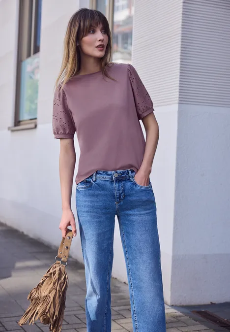 Eine Frau steht an einer Hauswand, trägt ein mauvefarbenes Top und blaue Jeans, hält eine Fransen-Tasche.
