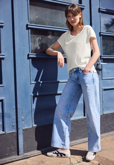 Een vrouw in wit T-shirt en losse jeans leunt nonchalant tegen een blauwe metalen deur buiten.