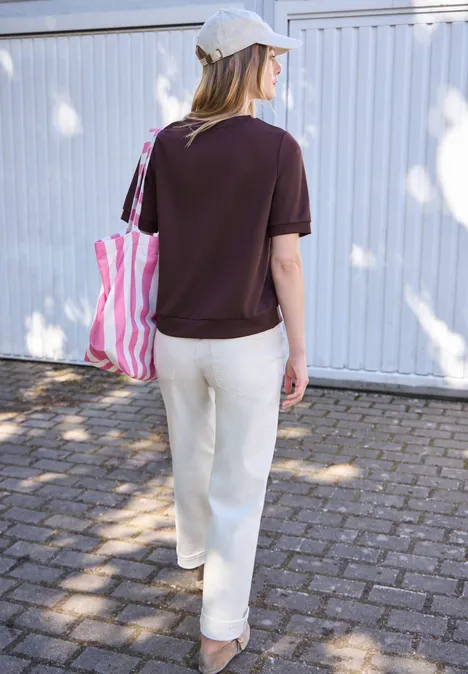 Basic T-Shirt mit Kurzarm und Rundhals very berry Frau von hinten mit weißer Kappe, dunkelrotem Shirt, weißer Hose und rosa-weiß gestreifter Tasche vor weißer Wand.