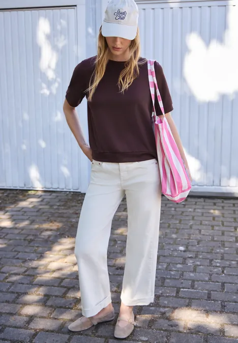 Basic T-Shirt mit Kurzarm und Rundhals very berry Frau in dunkelbraunem T-Shirt, weißer Hose, Schirmmütze und gestreifter Tragetasche steht vor heller Wand.