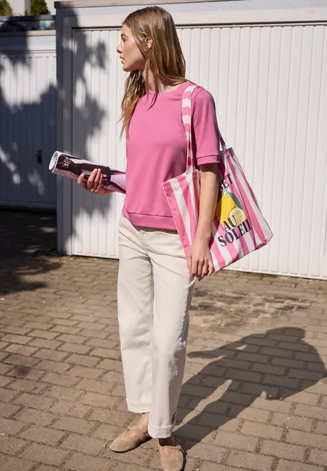 Vrouw in roze shirt en witte broek voor een wit hek, met telefoon in de hand en kleurrijke tas.