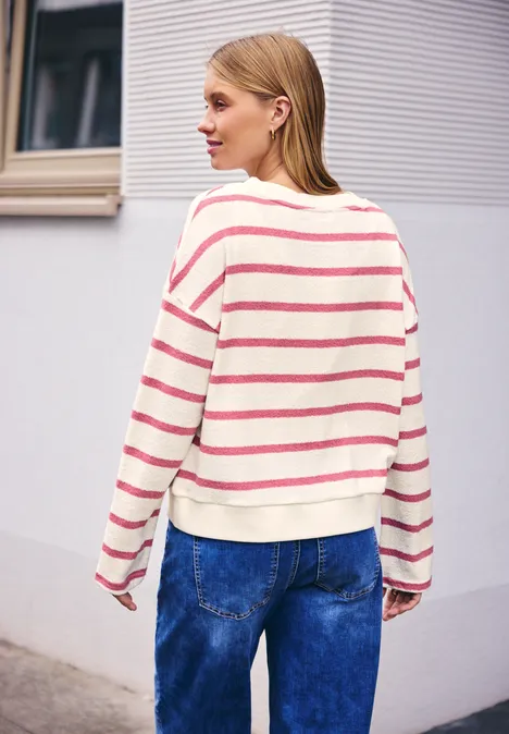Eine Person steht vor einer Wand, trägt einen gestreiften Pullover und blaue Jeans.