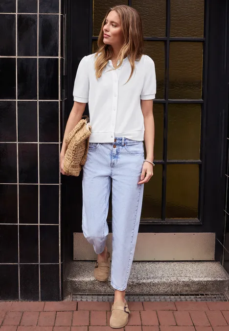 Femme en chemise blanche manches courtes, jean bleu clair et espadrilles beiges, sac en paille devant un portail carrelé noir.