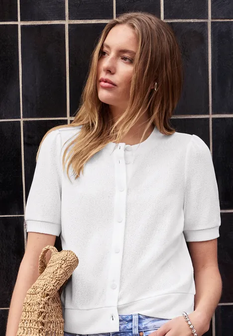 Femme en cardigan blanc à manches courtes, tenant un sac en osier devant un mur de carrelage sombre.