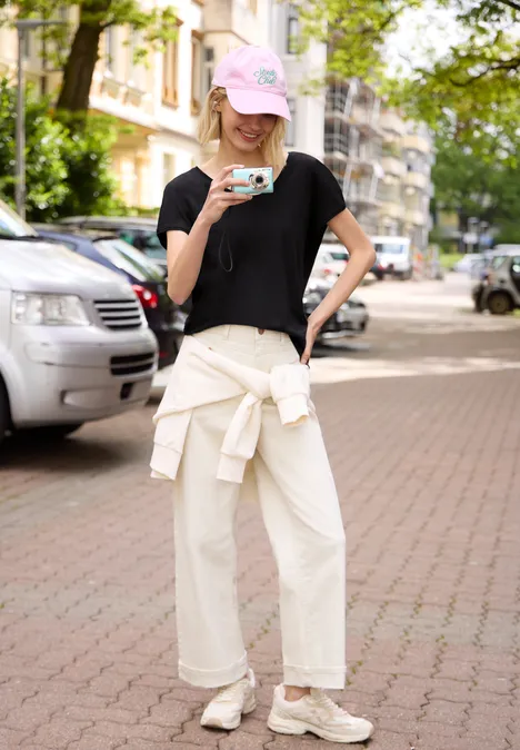 Junge Frau mit rosa Cap, schwarzem Shirt und weitem weißen Hose trinkt Kaffee auf Gehweg vor parkenden Autos.