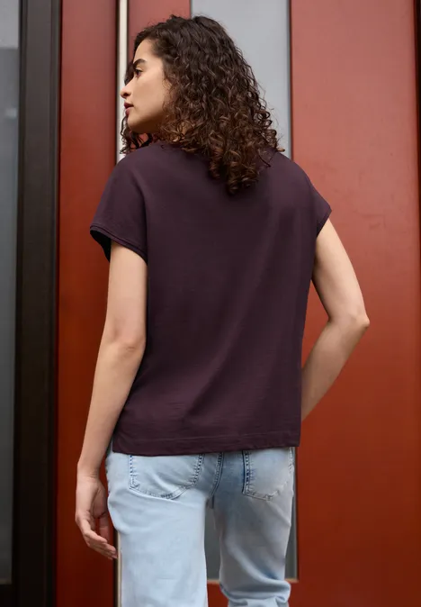 Dropped Shoulder Shirt aus Jersey very berry Frau mit lockigen Haaren steht mit dem Rücken zur Kamera, trägt dunkelviolettes Shirt und helle Jeans.