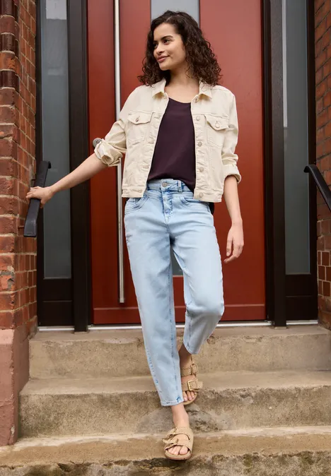 Dropped Shoulder Shirt aus Jersey very berry Frau in hellbeiger Jacke, schwarzem Top und hellblauen Jeans steht auf einer Treppe vor roter Tür.