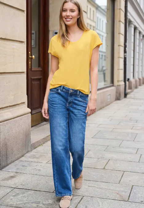 Vrouw in geel t-shirt en wijde jeans wandelt over de stoep langs historische gevels.