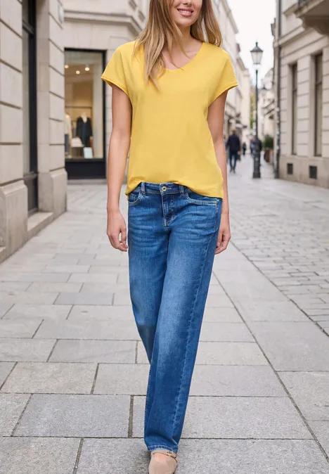 Een vrouw in een geel T-shirt en een blauwe spijkerbroek wandelt over een geplaveide straat.