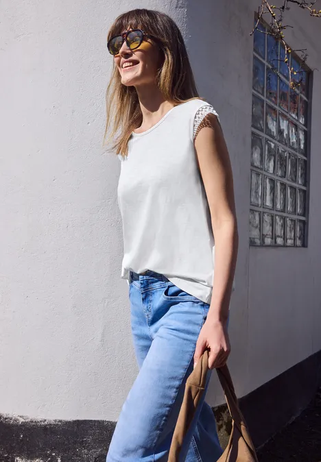 Vrouw in wit shirt en blauwe jeans leunt tegen een witte muur en houdt een bruine tas vast.