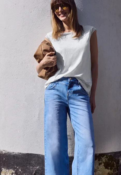 Vrouw in wit shirt en blauwe jeans voor een lichte muur, met een bruine jas in haar hand.